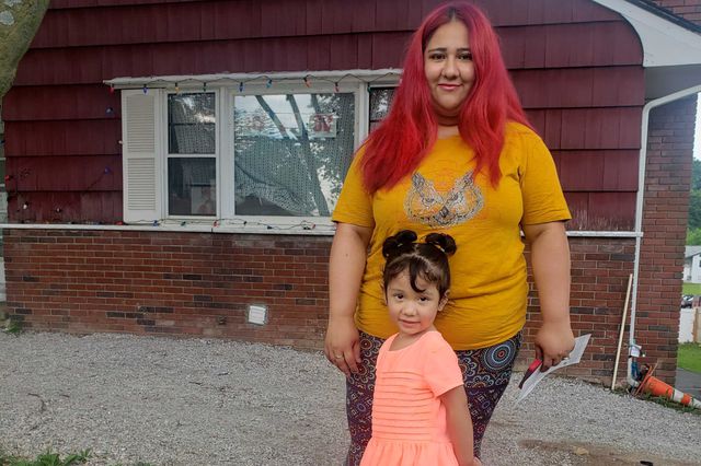 Ana Maeda Gonzalez has red hair and is wearing a yellow t-shirt and her young daughter stands in front of her, wearing a pink dress. They are standing in front of a red house.